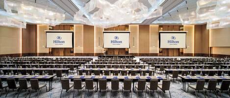 A large conference room with rows of tables and chairs, three large screens displaying the Hilton Kuala Lumpur logo, and a modern ceiling design.