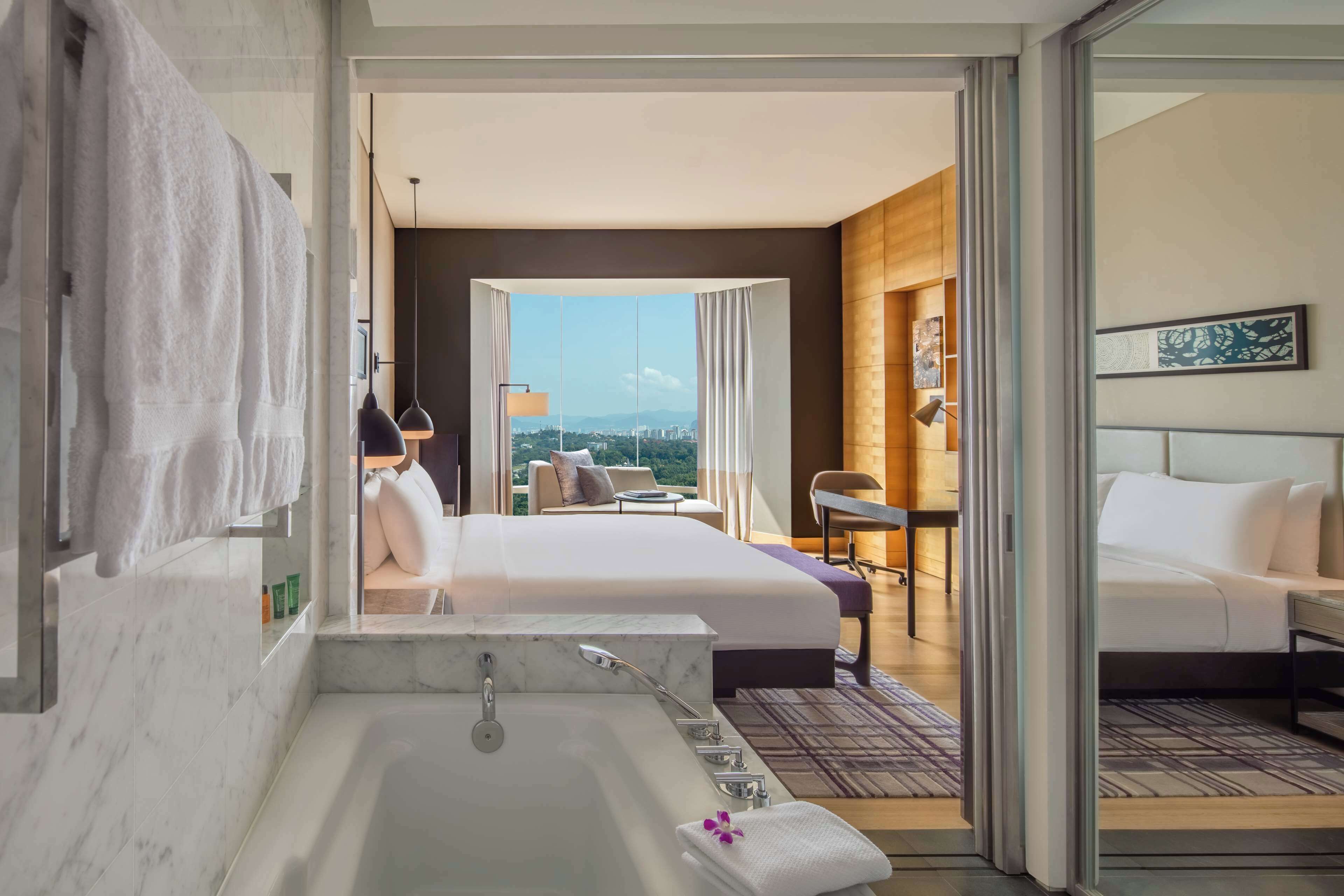 Una habitación de hotel moderna con una cama grande, un área de estar separada, un baño con bañera y vistas a la ciudad.