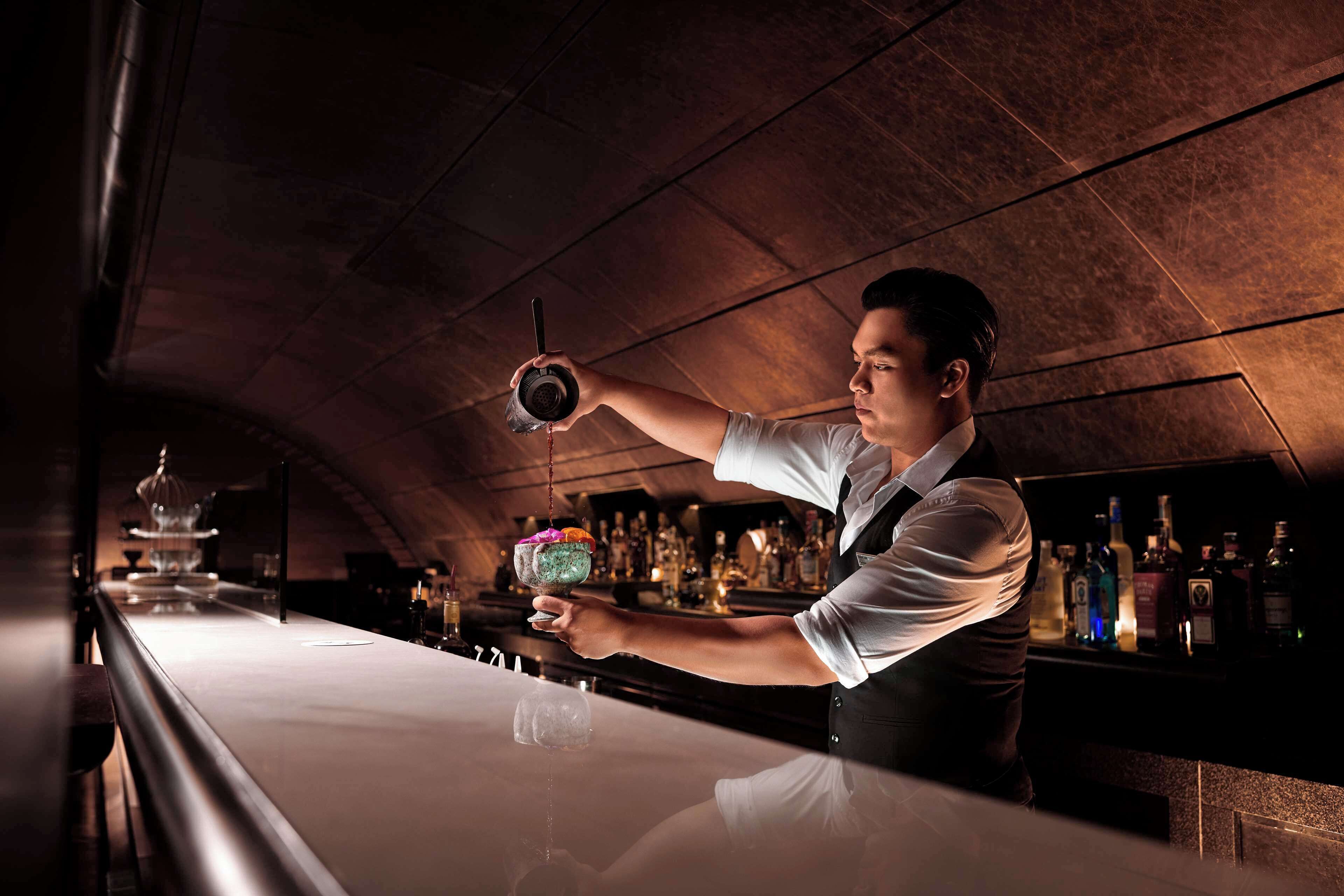 A bartender preparing a cocktail at a bar with a polished countertop and a selection of bottles in the background.