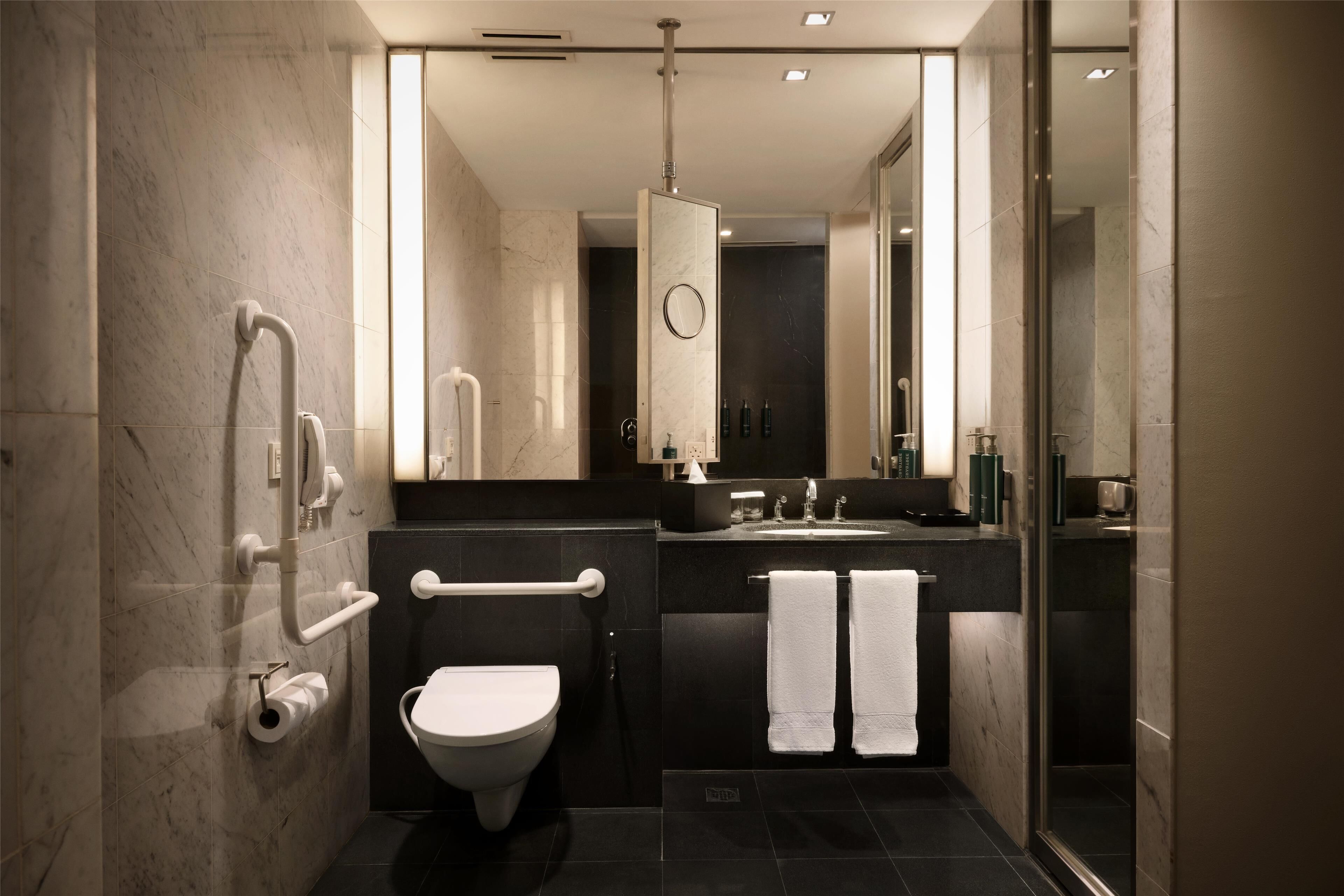 A modern bathroom with a toilet, bidet, and shower. It features a large mirror, dark tiles, and a glass shower enclosure.