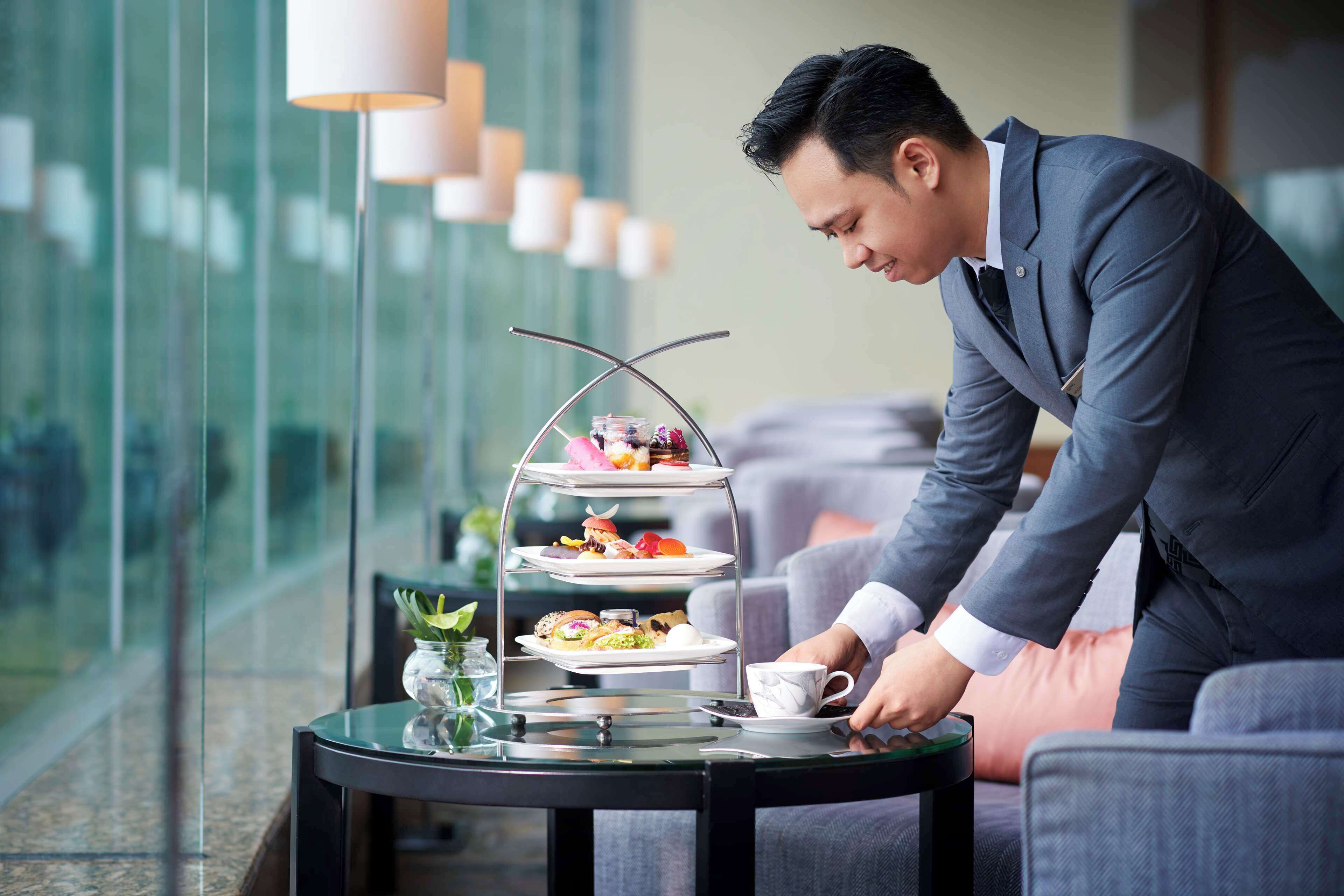 Un homme en costume est en train de disposer un présentoir à trois niveaux rempli de desserts sur une table dans le hall d’un hôtel moderne, doté de grandes fenêtres et de meubles contemporains.