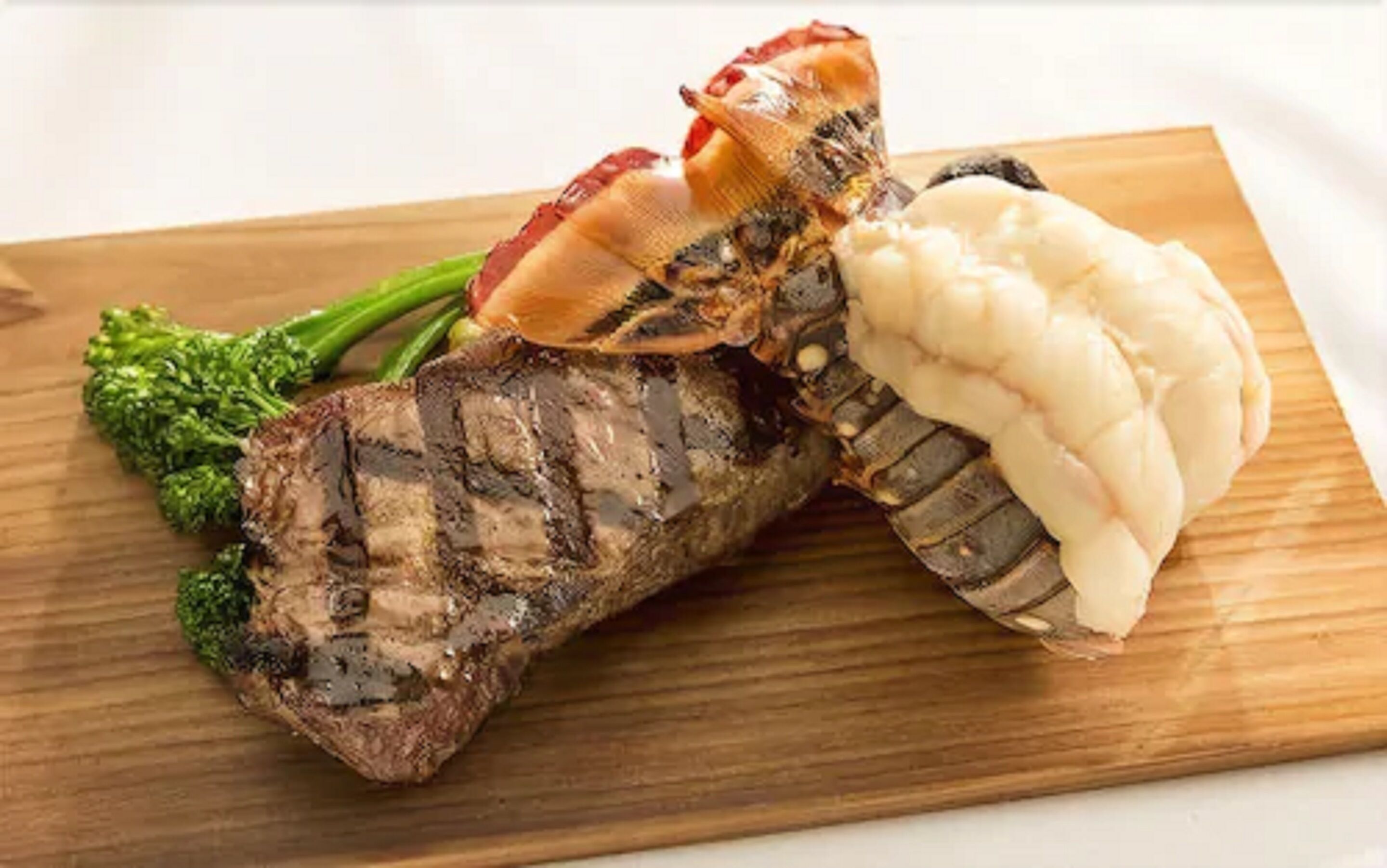 A steak with a lobster tail and broccoli on a wooden board.