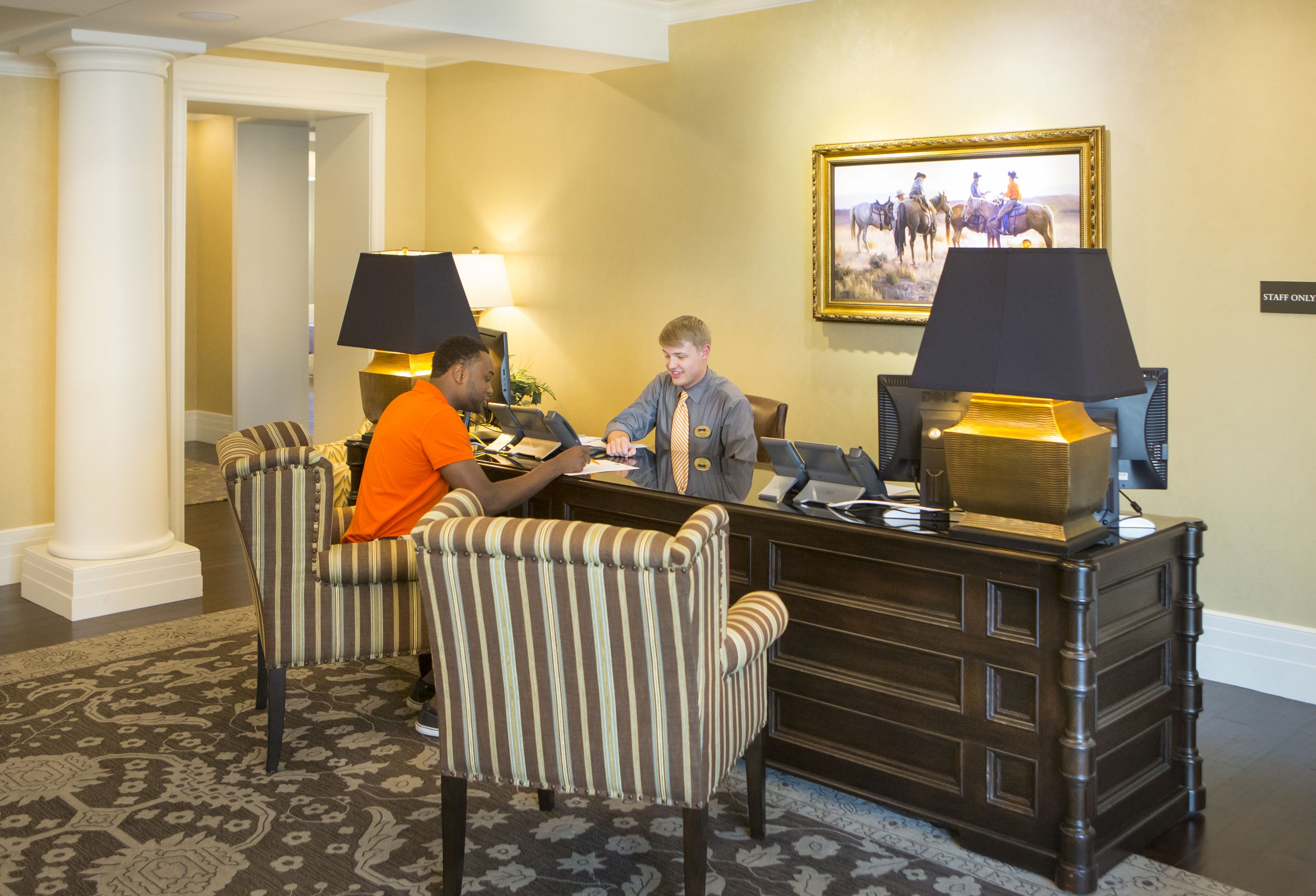 A reception area with two staff members seated at a desk, a framed picture on the wall, and a patterned carpet.