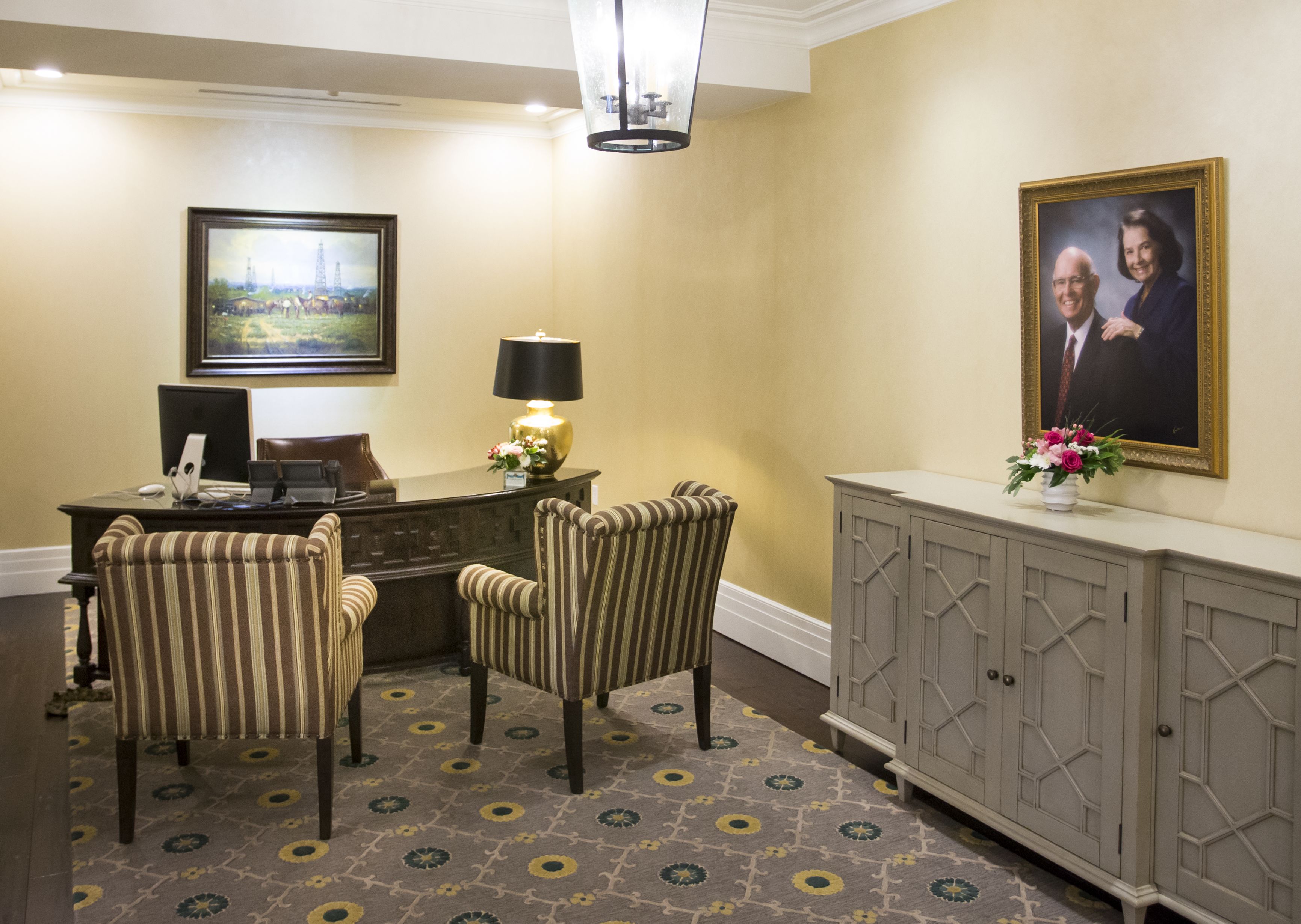 A reception area with a desk, two striped chairs, a lamp, a framed portrait, and a patterned carpet.