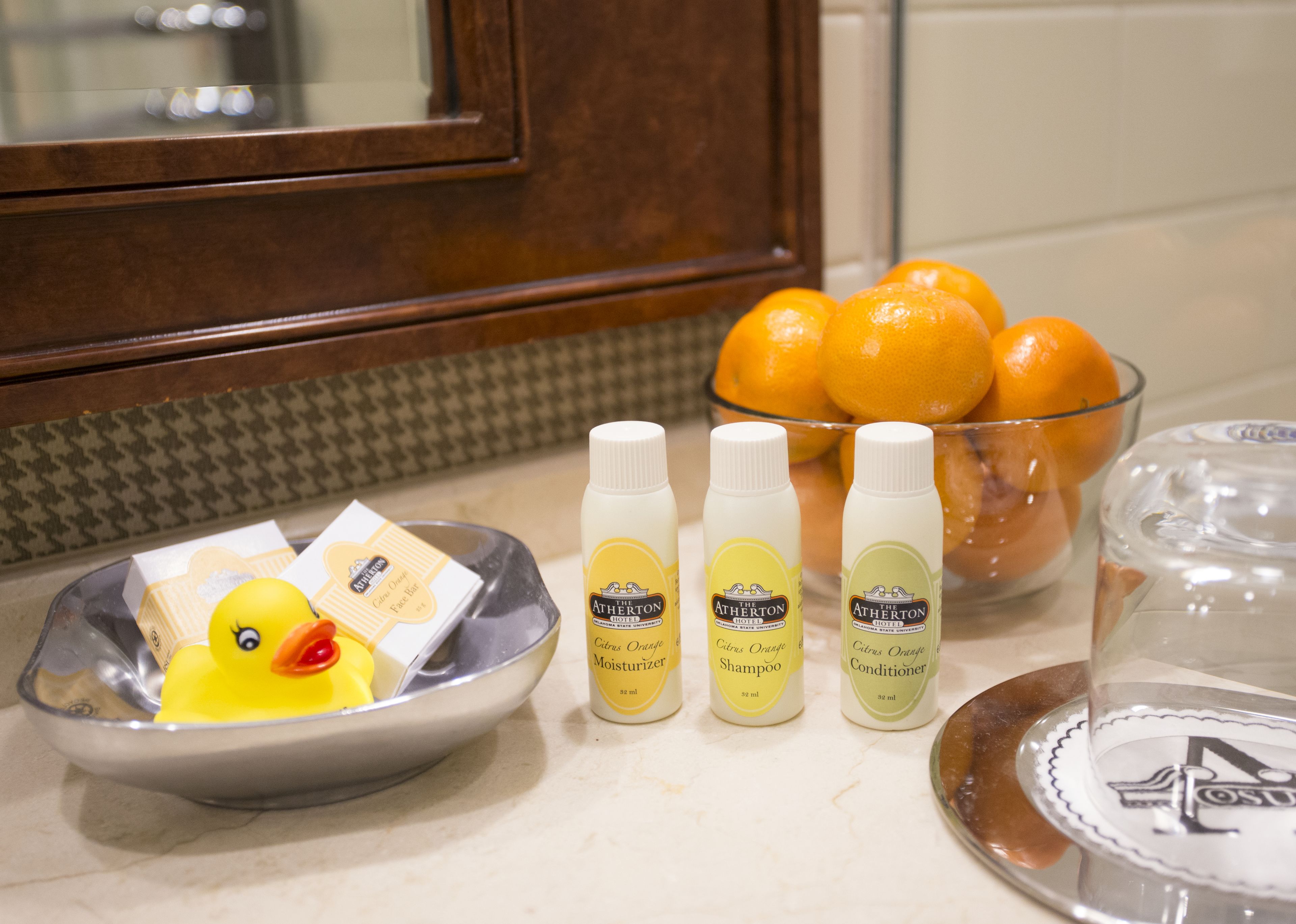 A kitchen counter with a bowl of oranges, a rubber duck, and three bottles of The Atherton brand personal care products.