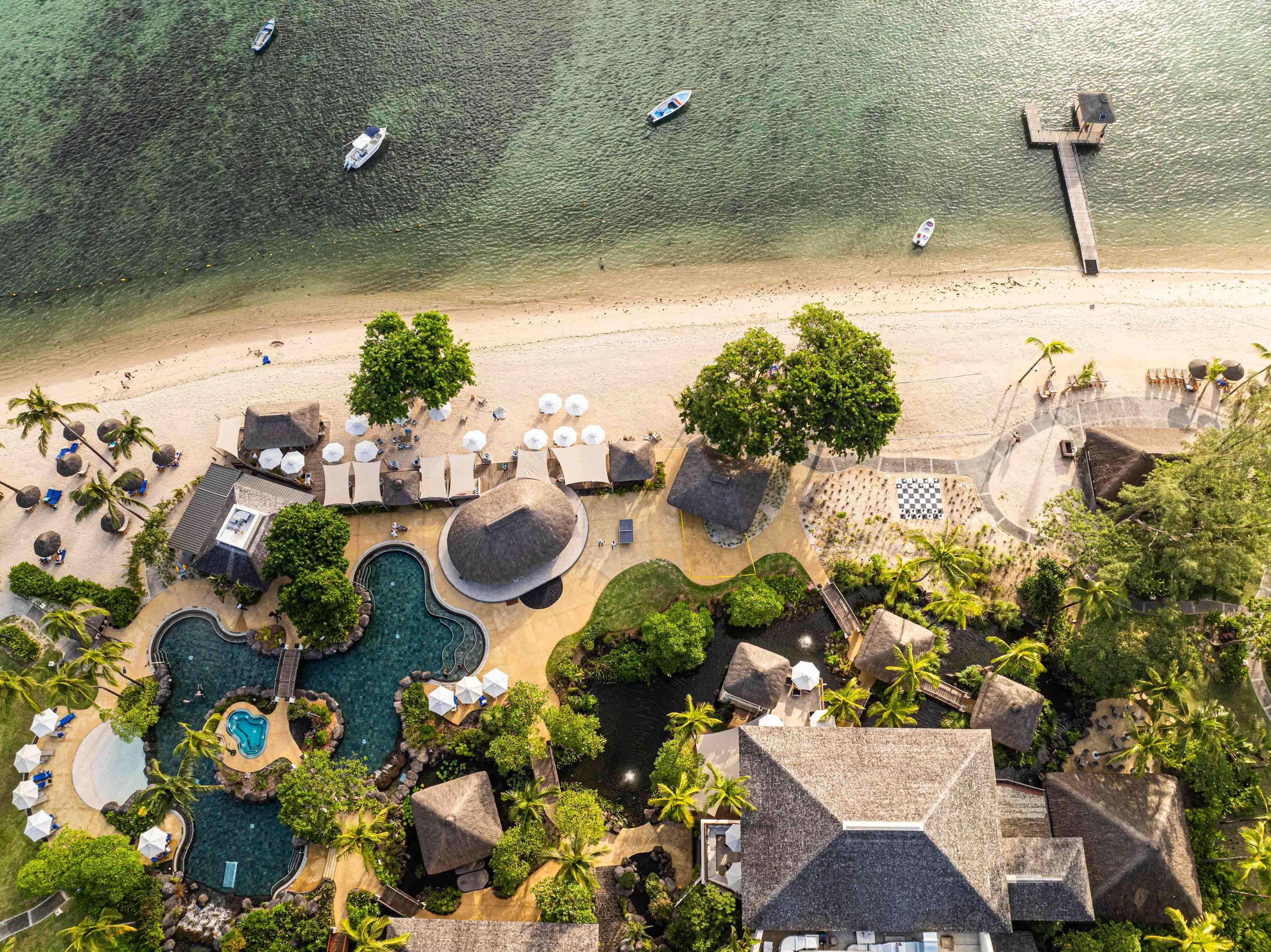 Vista aérea de um resort com praia, piscina e construções com telhado de palha.