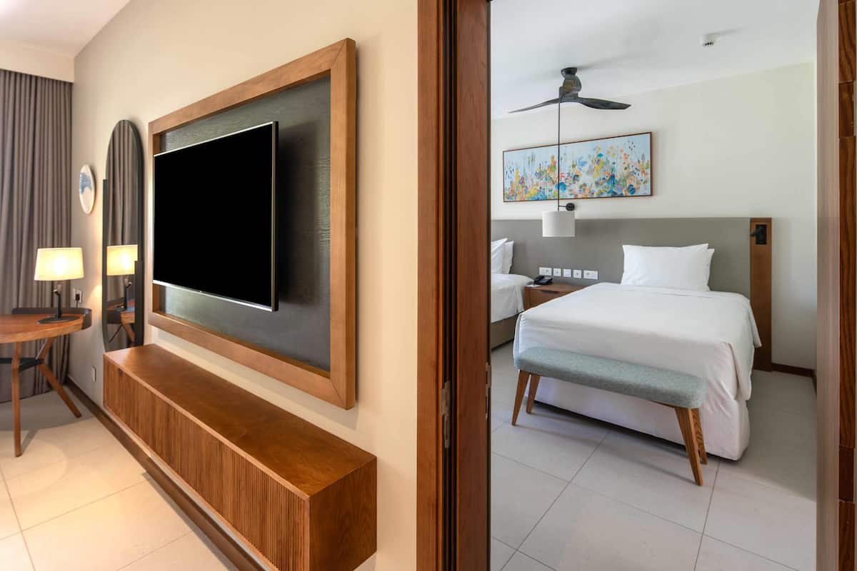 Une chambre d’hôtel dotée d’un grand téléviseur à écran plat fixé au mur, d’une console en bois, d’un lit avec un banc gris, d’un ventilateur de plafond et d’un tableau accroché au mur.
