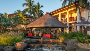 Ein Resort mit einem strohgedeckten Pavillon, einem Pool mit Wasserfall und einem mehrstöckigen Gebäude mit Balkonen.