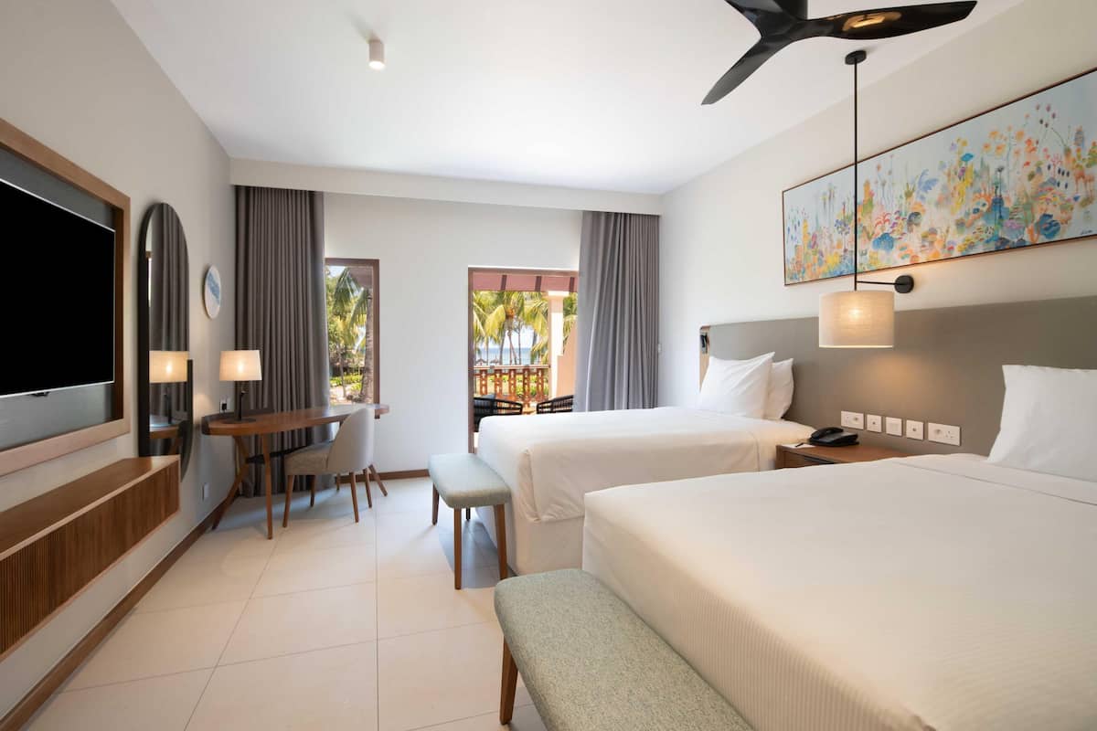 Une chambre d’hôtel équipée d’une télévision, de deux lits, d’un bureau avec une chaise, d’un banc, d’un ventilateur de plafond et d’un tableau au mur.