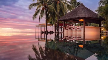 An infinity pool with a thatched-roof structure, palm trees, and a sunset view.