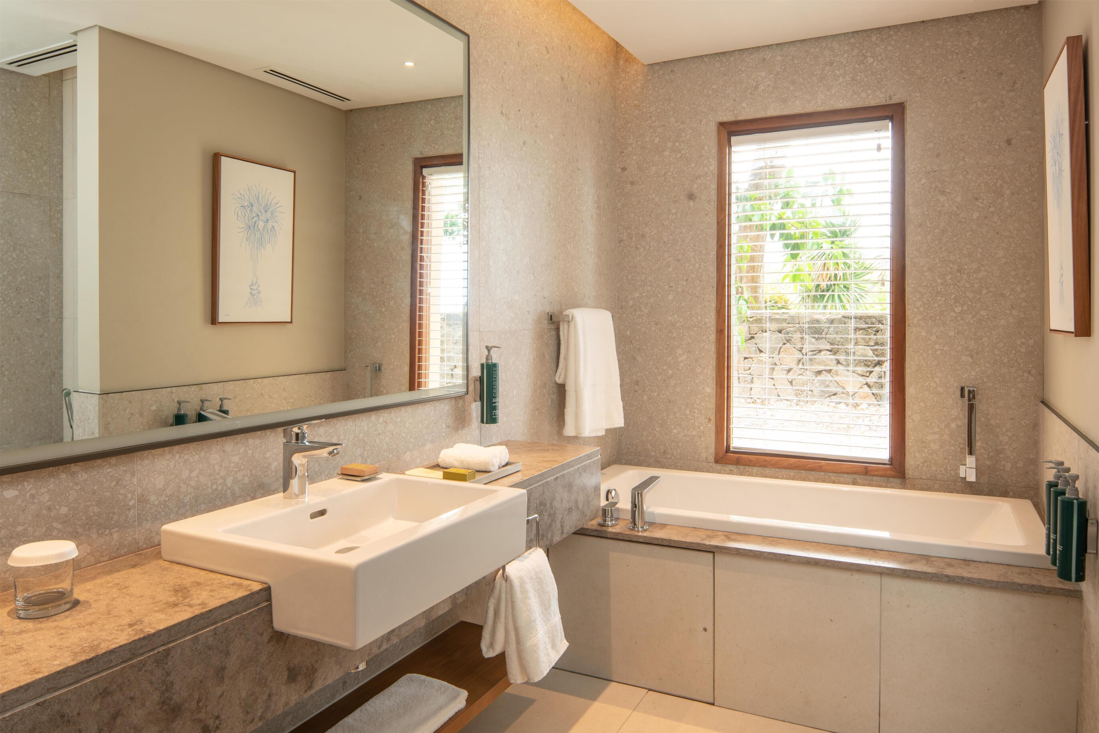Un baño moderno con un espejo grande, dos lavamanos, una bañera y una ventana con persianas.