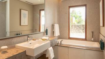 Un baño moderno con un espejo grande, dos lavamanos, una bañera y una ventana con persianas.