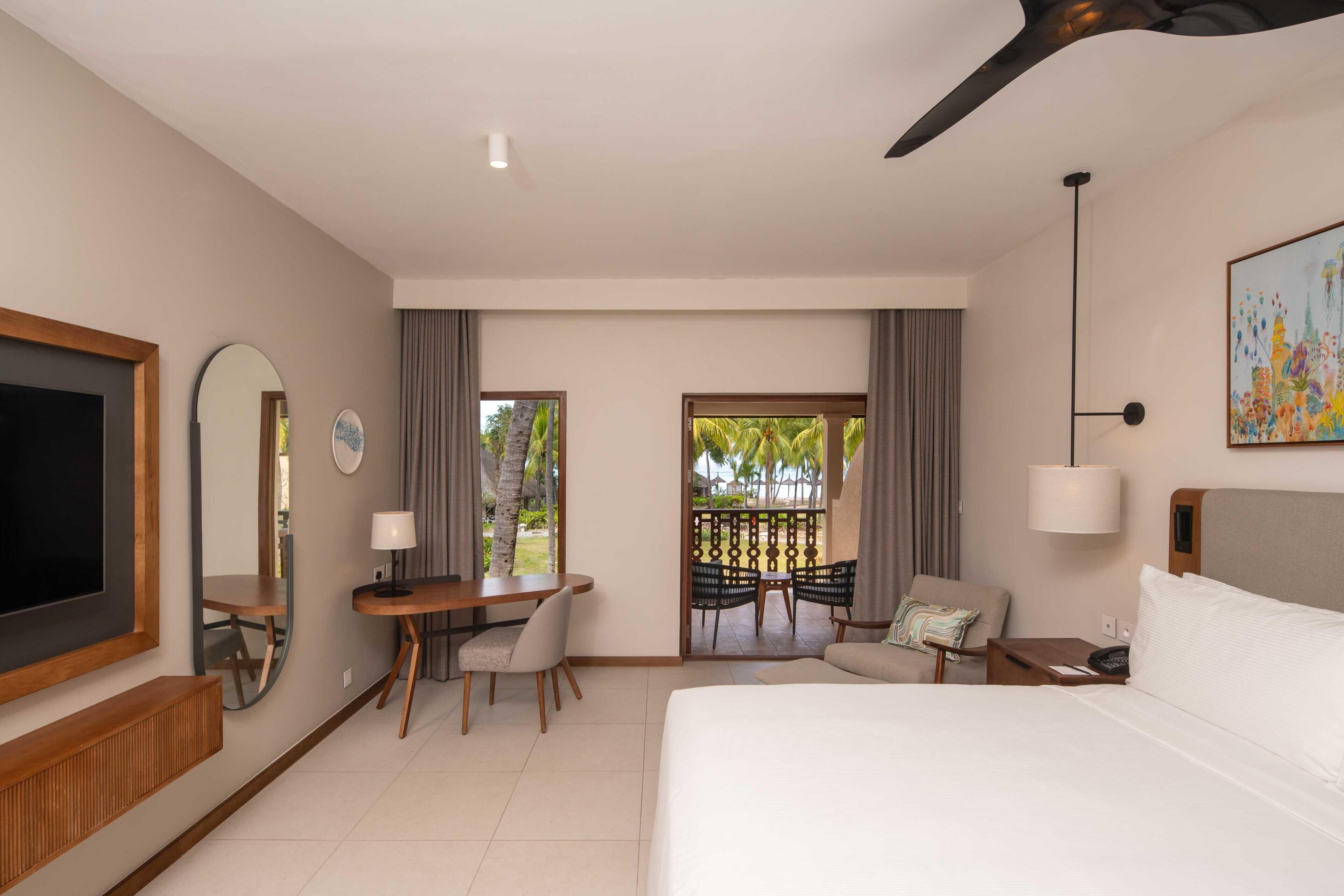 Habitación de hotel con cama, escritorio, sillas, televisor, ventilador de techo y balcón con vista a palmeras.