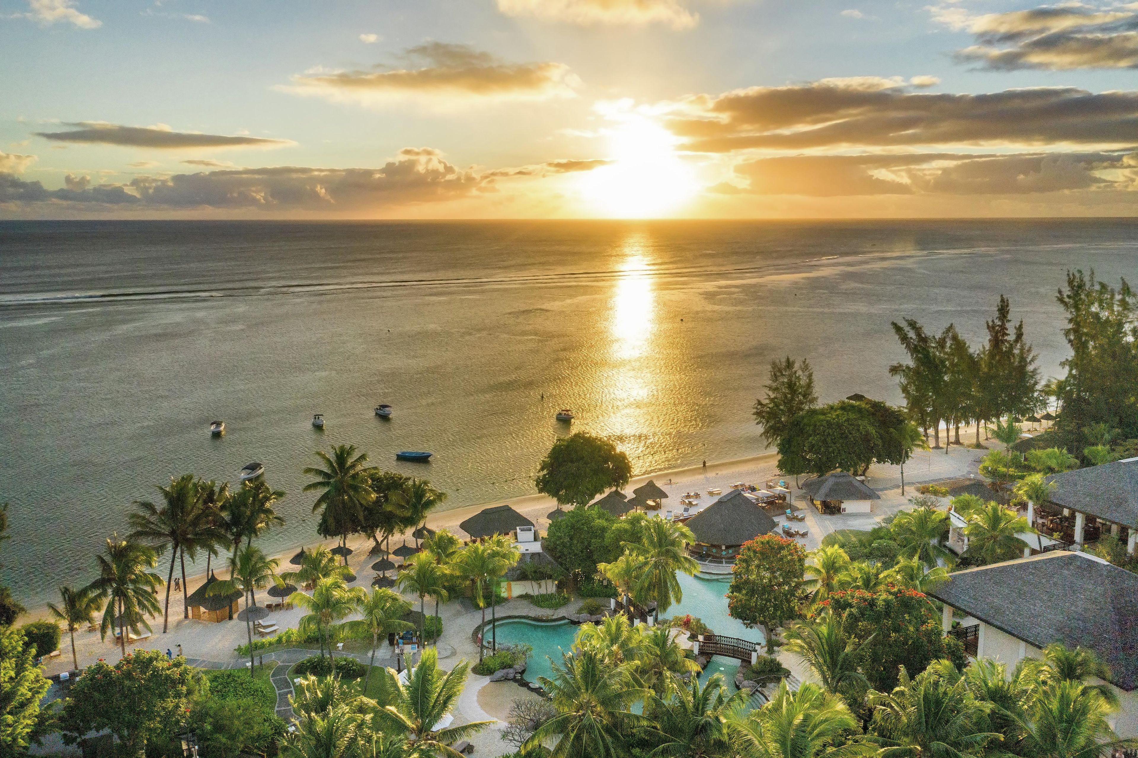 A resort with a swimming pool, palm trees, and a view of the ocean at sunset.