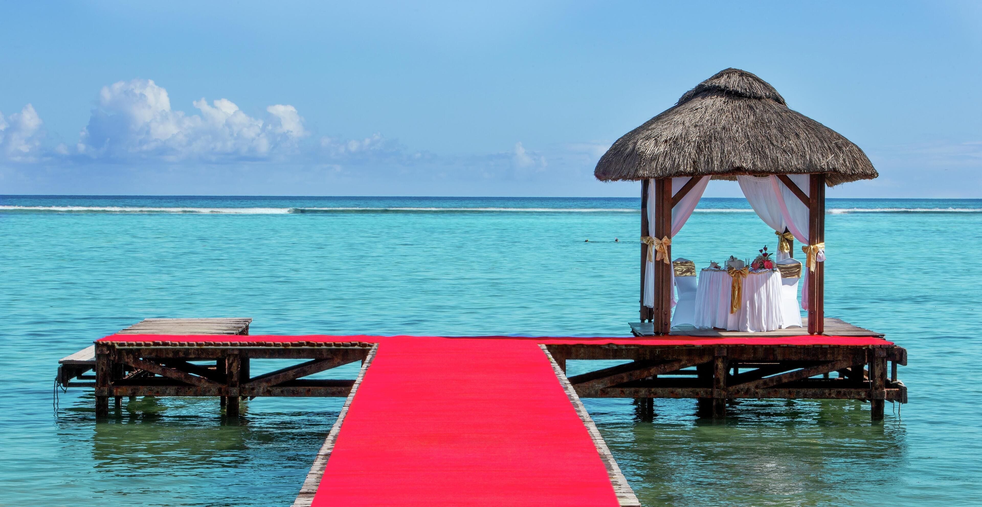 A wooden pier with a red carpet leading to a thatched-roof structure with white drapery and gold accents.