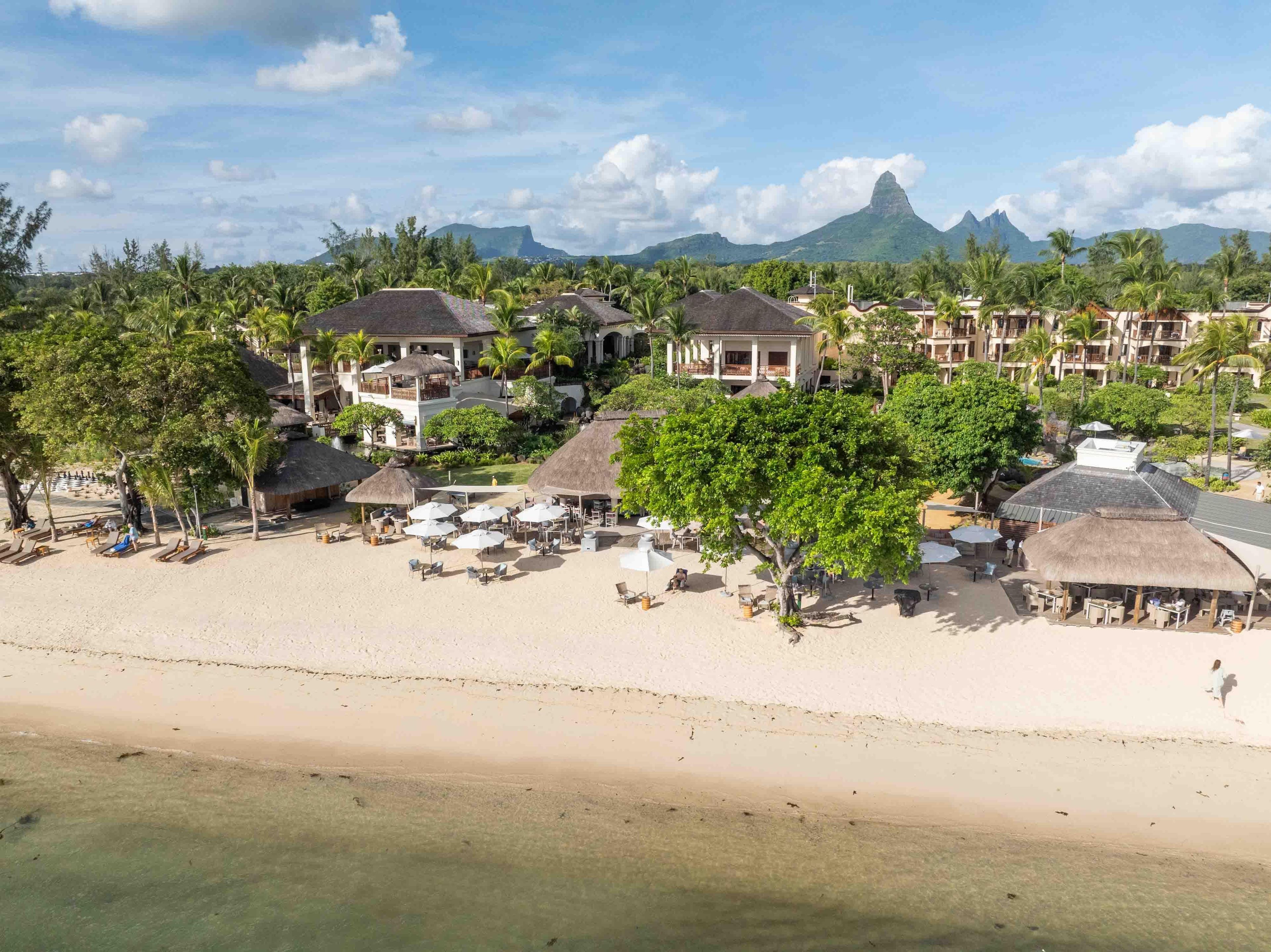 Un complejo turístico con varias construcciones, una zona de playa con sombrillas y tumbonas, y montañas al fondo.
