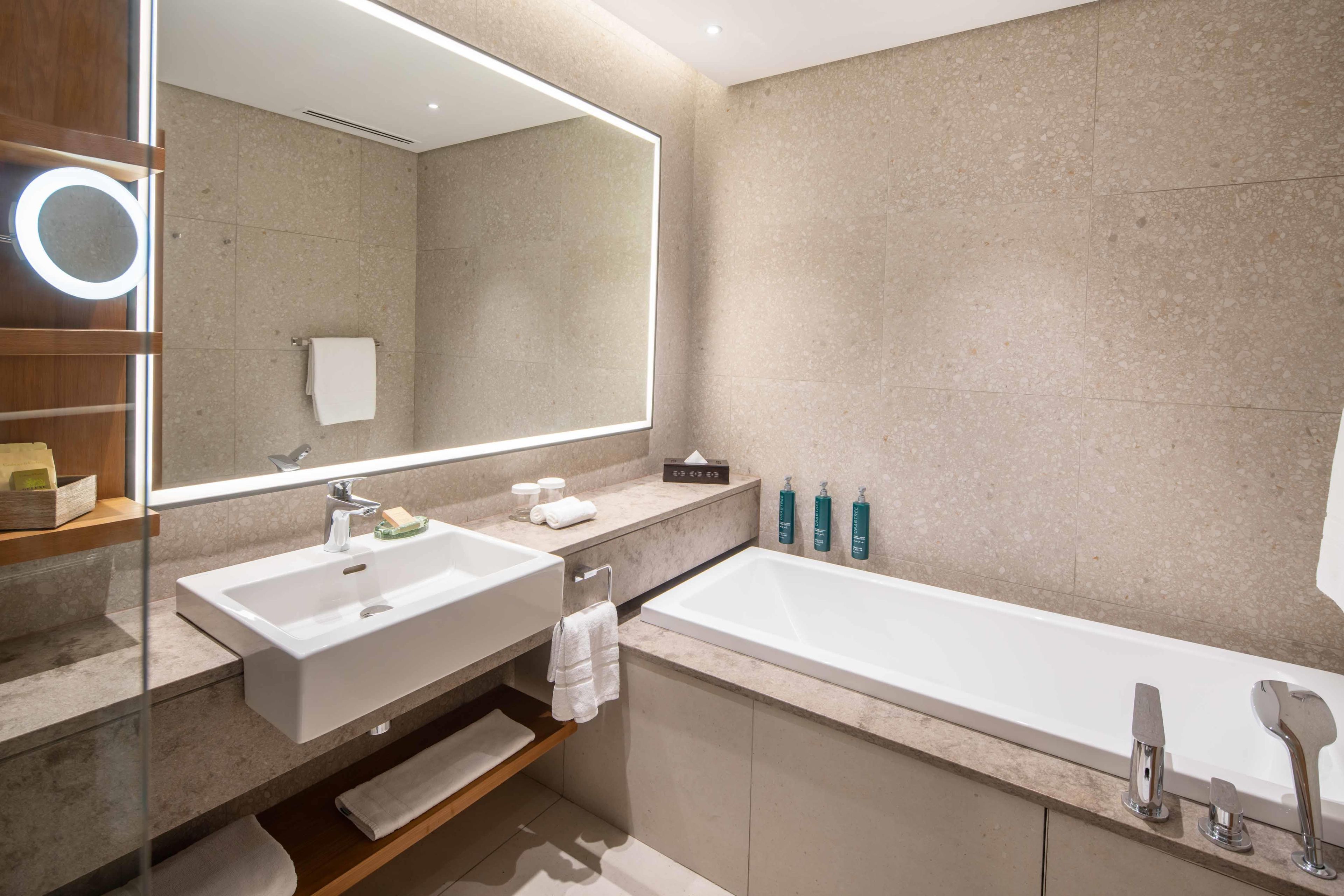 Un baño moderno con un espejo grande, dos lavamanos, una bañera y un estante con artículos de aseo.