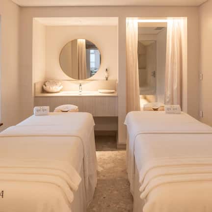 A hotel room with two beds labeled 'THE SPA,' a round mirror, a sink, and a shower area.