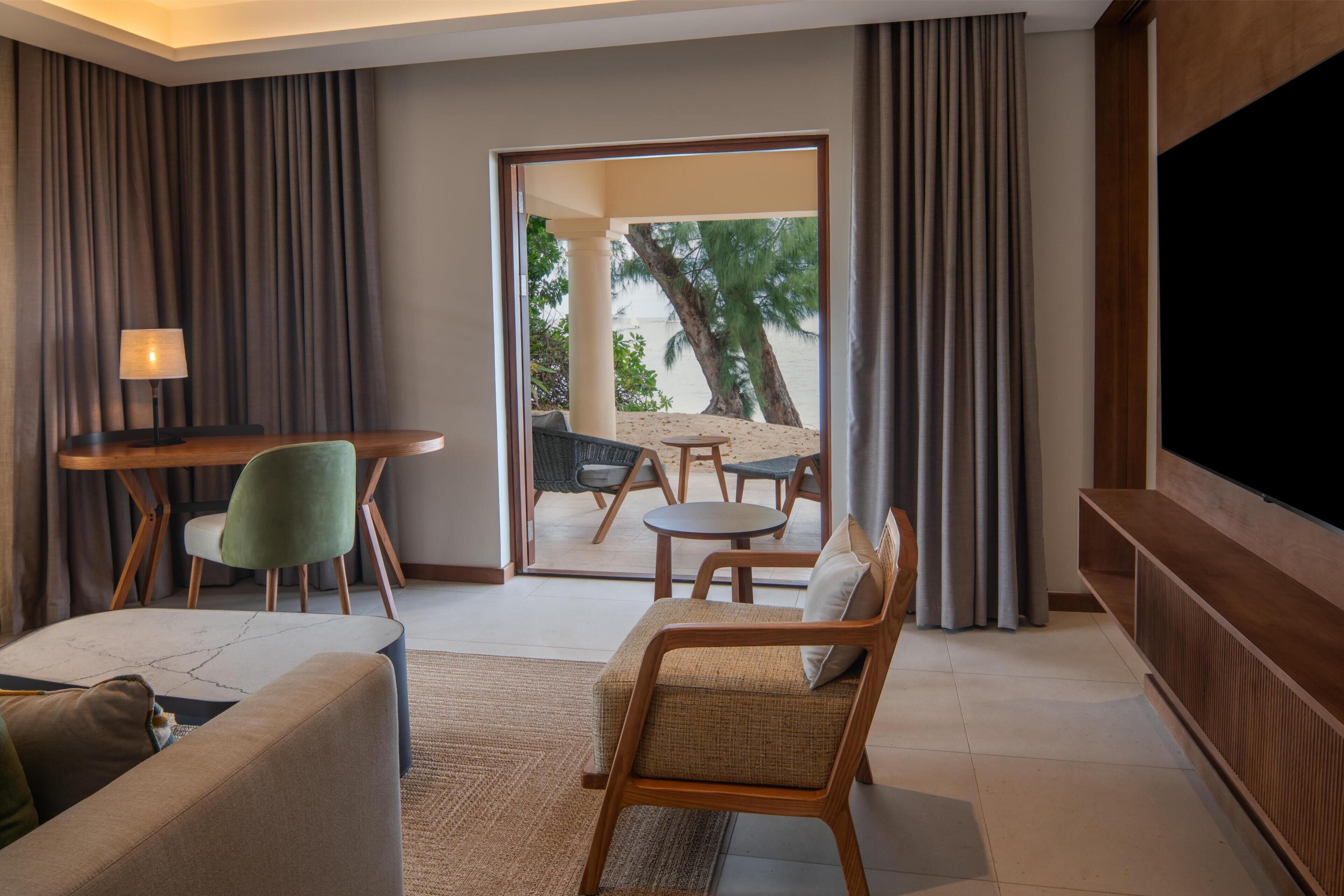 A modern hotel room with a sofa, a dining table with chairs, a TV, and a view of the outdoors.