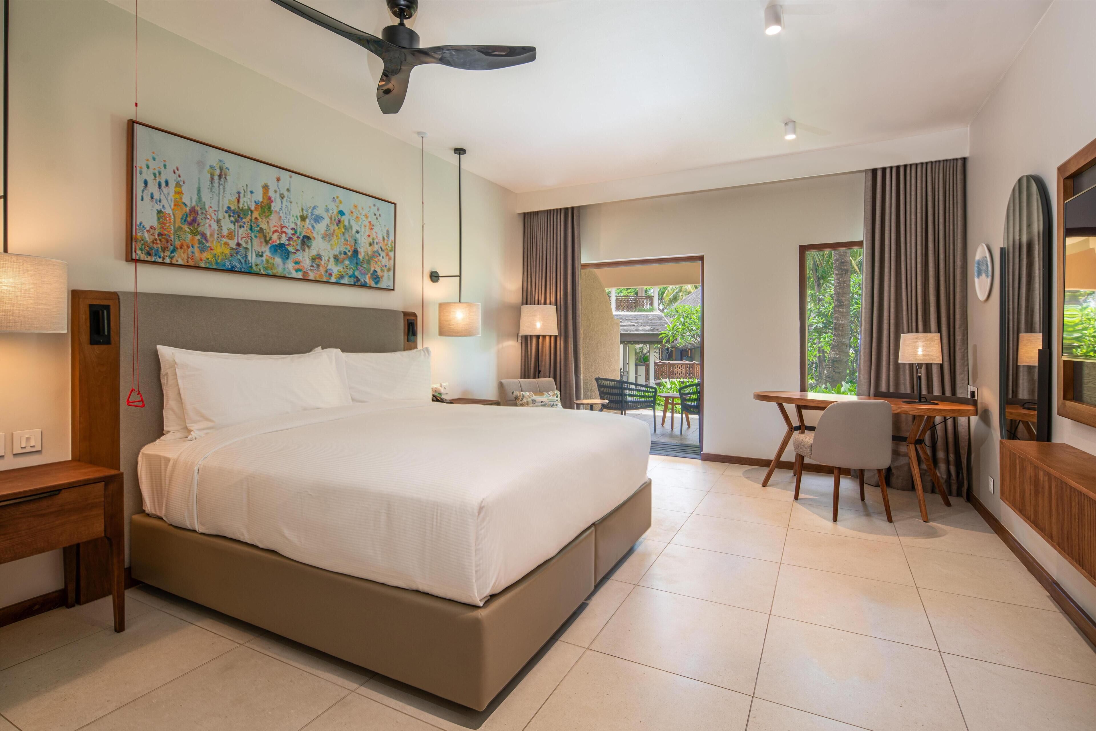 A hotel room with a bed, desk, chair, ceiling fan, and artwork on the wall.