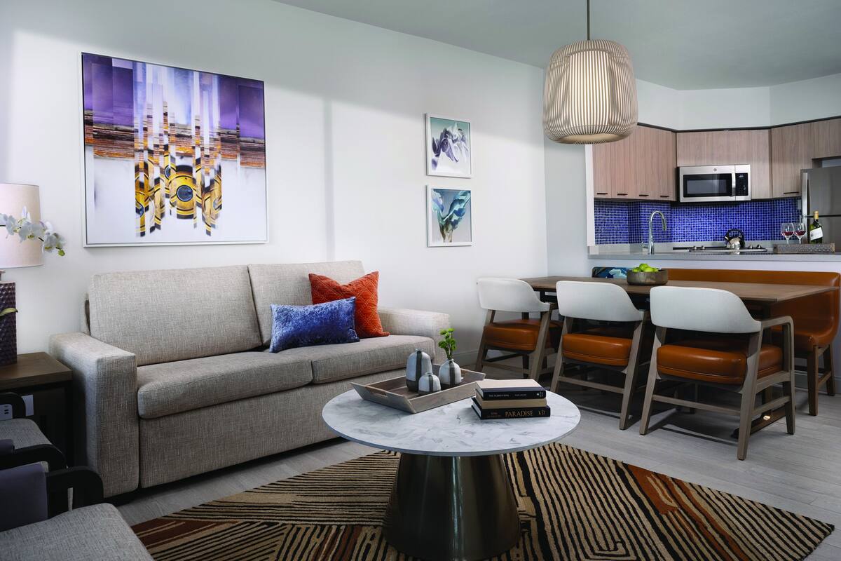 Ein modernes Wohnzimmer mit einer Couch, einem Couchtisch und Kunstwerken. Im Hintergrund ist eine Küche mit Mikrowelle und Spüle zu sehen.
