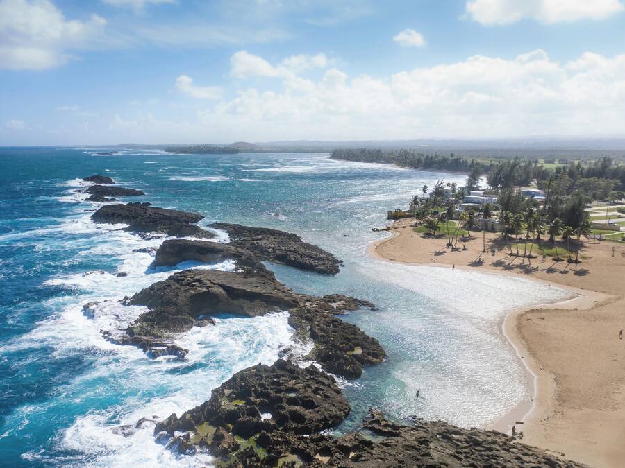 A coastal view with rocky formations, a sandy beach, and a resort area with buildings and trees.