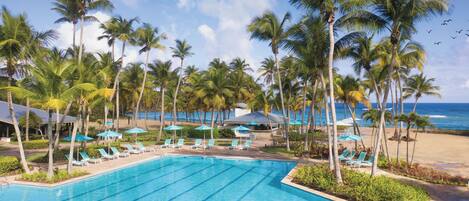 A swimming pool with lounge chairs and umbrellas, surrounded by palm trees and a beach view.