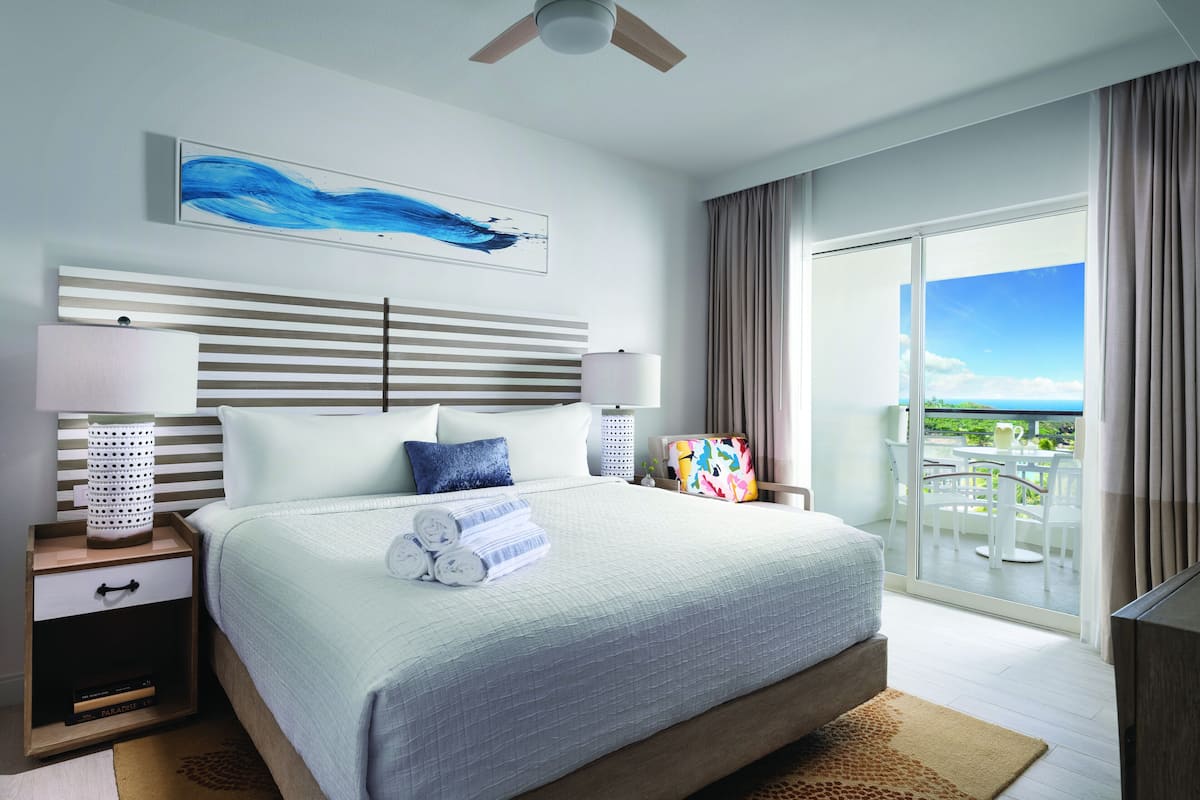 Ein modernes Hotelzimmer mit einem großen Bett, einem Deckenventilator, Blick auf den Ozean und einem Balkon mit Gartenmöbeln.