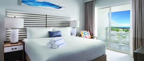 Habitación de hotel moderna con una cama grande, ventilador de techo, vista al océano y balcón con mobiliario exterior.