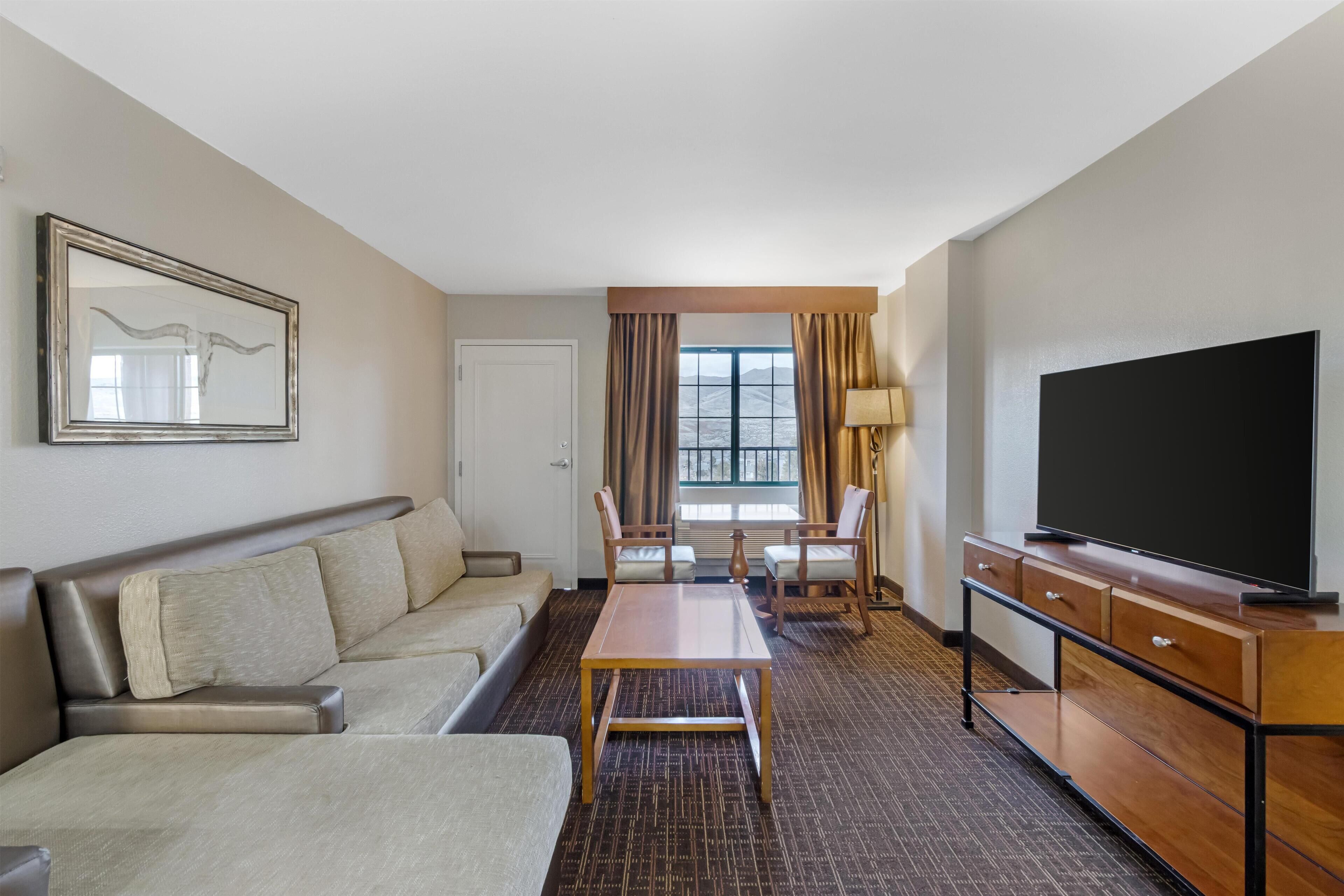 A hotel room with a sofa, a table, chairs, a TV, and a window with curtains.