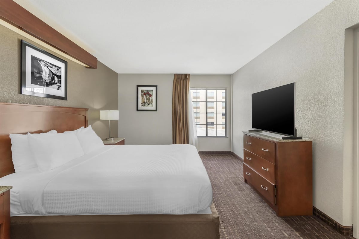 A hotel room with a bed, a TV, a dresser, a window with curtains, and framed pictures on the wall.