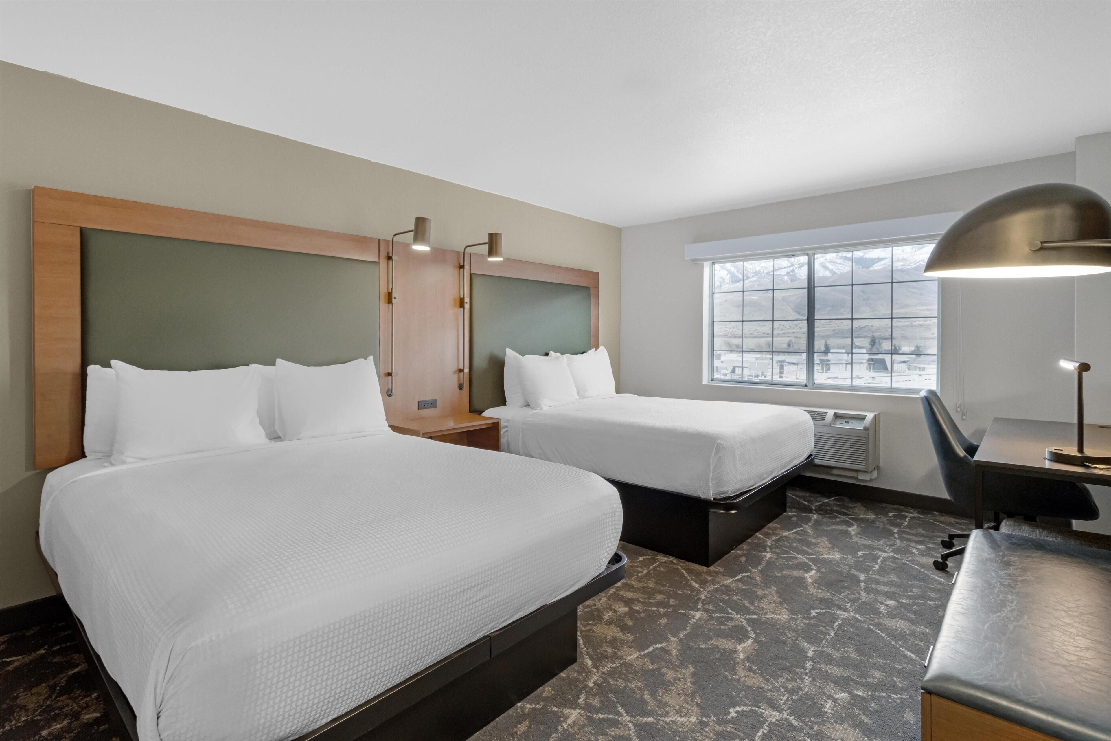 A hotel room with two beds, a desk, a chair, a lamp, and a large window with a view.
