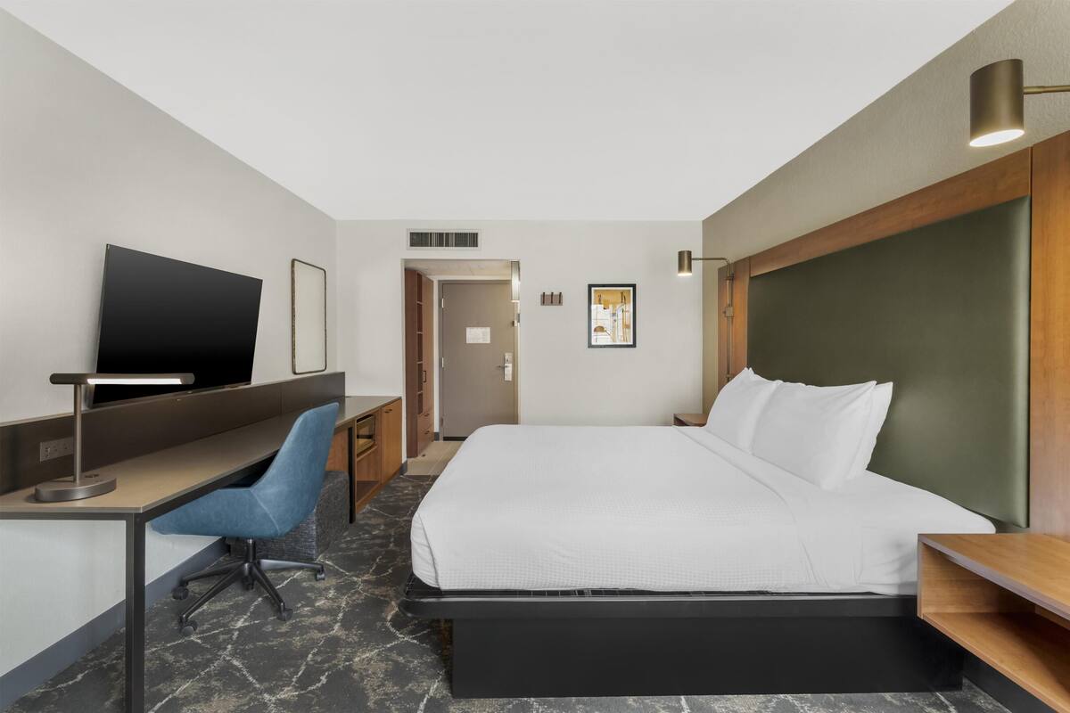 A hotel room with a bed, desk, chair, TV, and a door leading to another room.