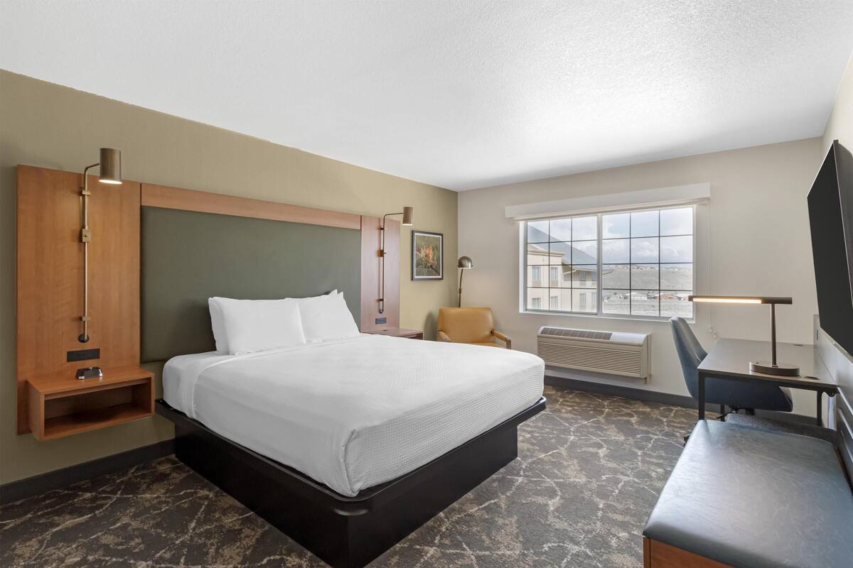 A hotel room with a bed, a desk, a chair, a TV, a window, and a wall-mounted shelf.