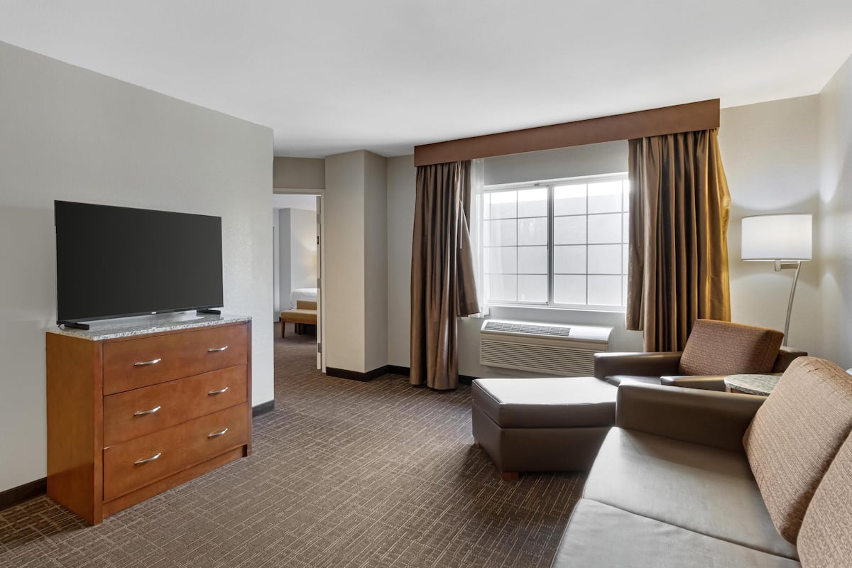A hotel room with a flat-screen TV on a wooden dresser, a sofa, a window with curtains, and a lamp.