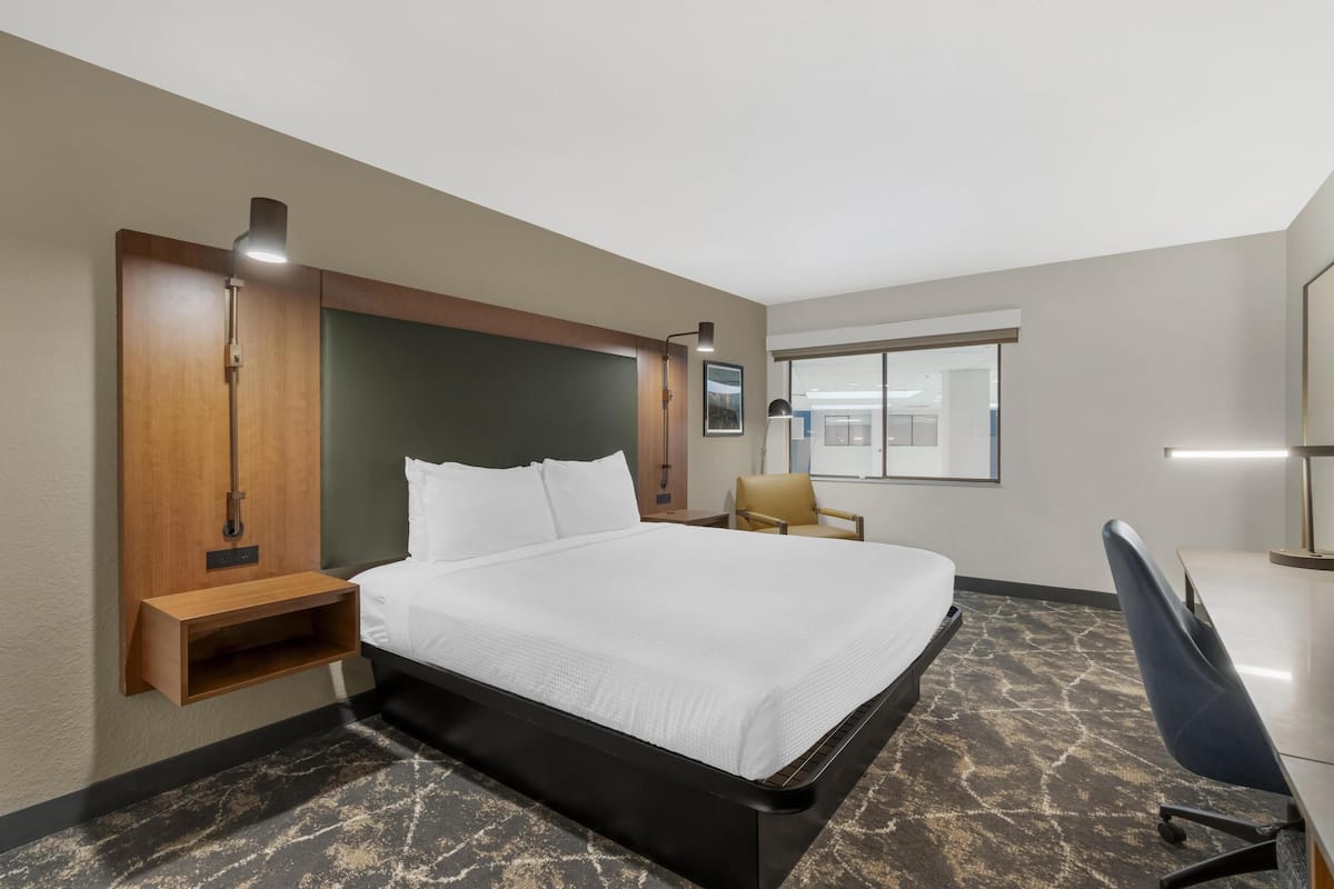 A hotel room with a bed, a desk, a chair, a nightstand, a lamp, and a window.