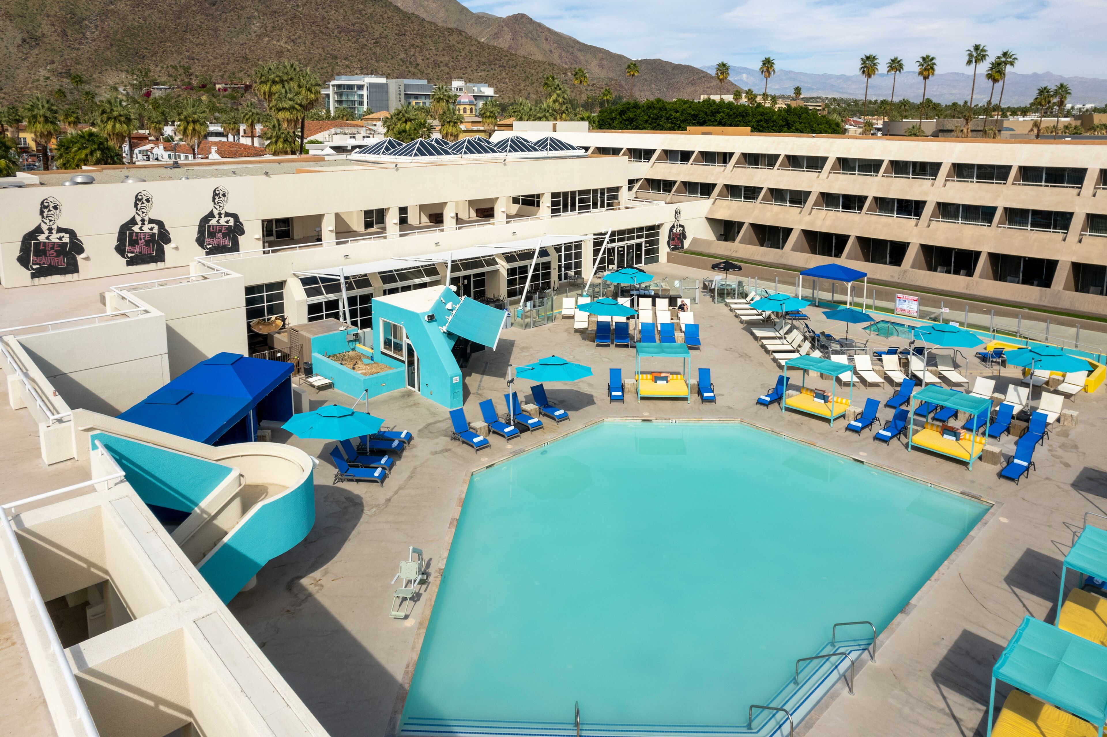 A pool area with multiple sun loungers and umbrellas, a large swimming pool, and a building with a distinctive architectural design.