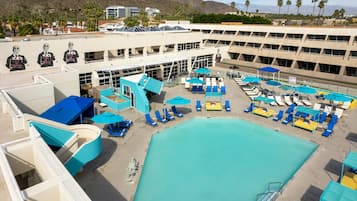 A pool area with multiple sun loungers and umbrellas, a large swimming pool, and a building with a distinctive architectural design.