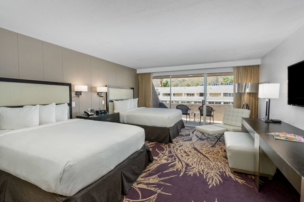 A hotel room with two beds, a desk, a chair, and a balcony with a view of buildings and trees.