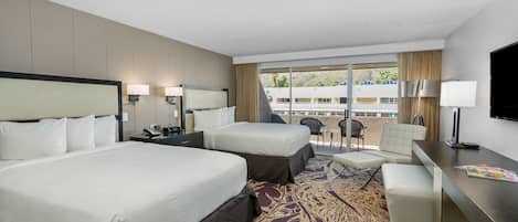 A hotel room with two beds, a desk, a chair, and a balcony with a view of buildings and trees.