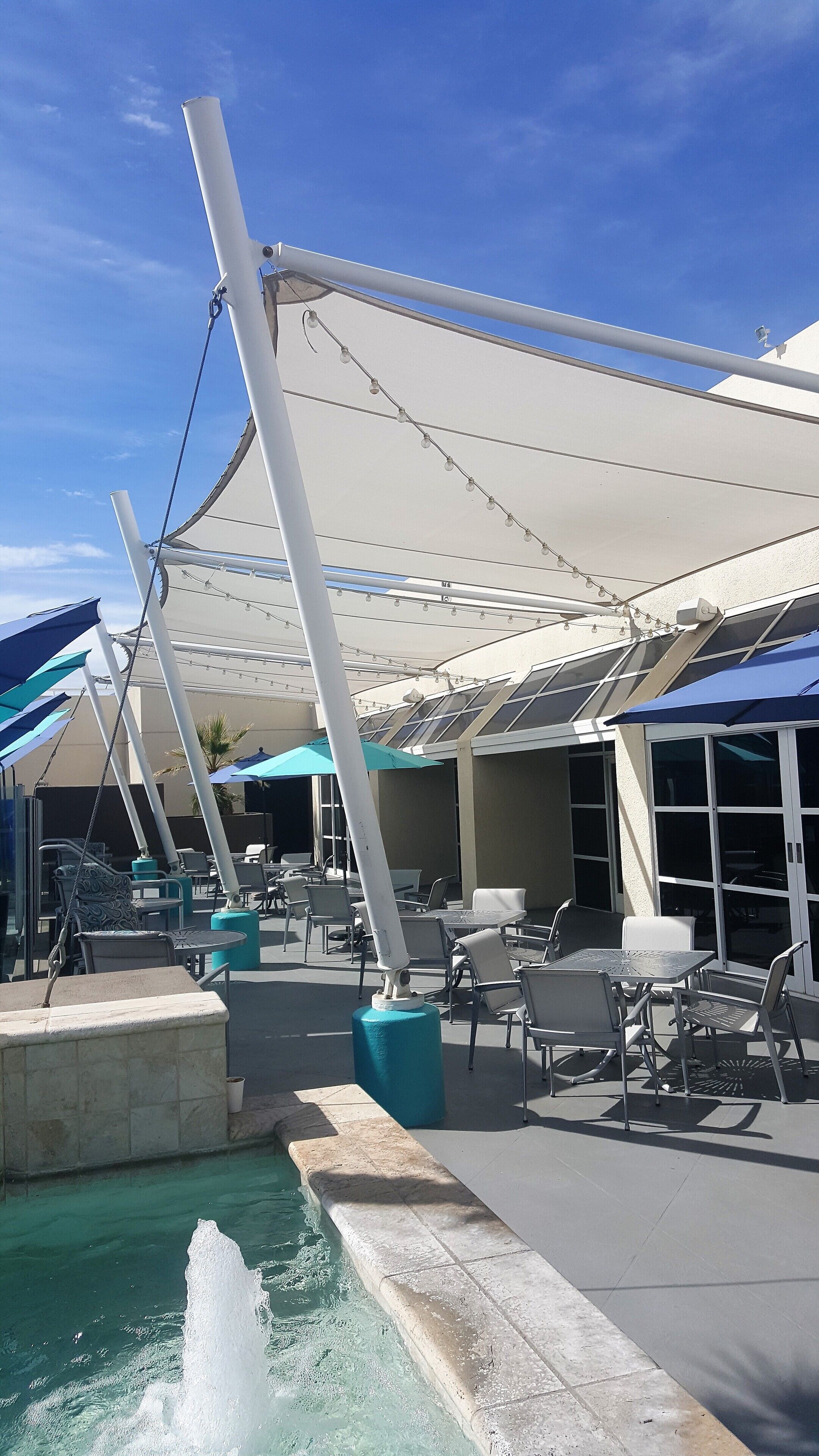 An outdoor seating area with tables and chairs, a pool with a water feature, and a covered patio area with umbrellas.