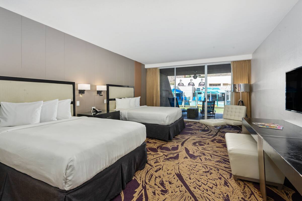 A hotel room with two beds, a desk, a chair, and a view of the outdoor area.