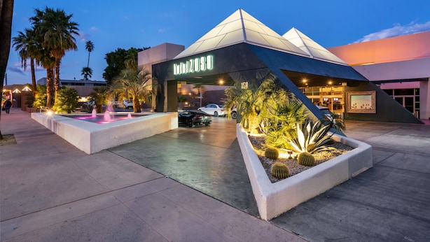 A modern building with a unique geometric design, a well-lit entrance, and a landscaped area with palm trees and a water feature. - Hotel Zoso (Palm Springs)