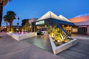 Un edificio moderno con un diseño geométrico único, una entrada bien iluminada y un área ajardinada con palmeras y una fuente de agua.