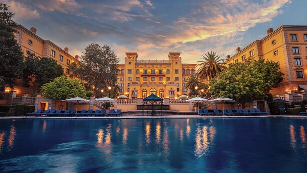 A hotel with a large pool, sun loungers, umbrellas, and a grand building with a central entrance. - Palazzo Hotel (Sandton)