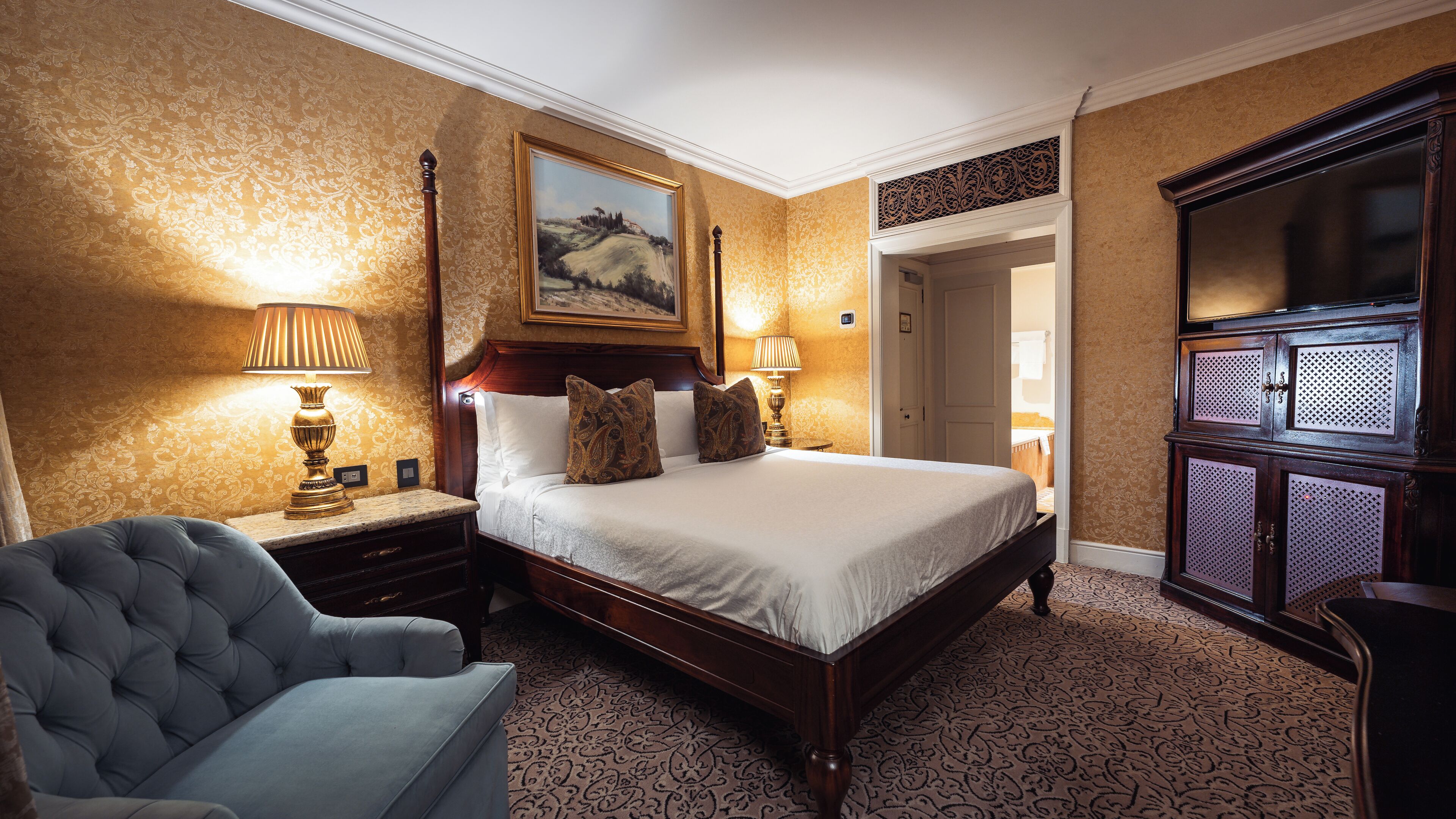 A hotel room with a large bed, two bedside lamps, a tufted armchair, a painting above the bed, and a wardrobe with a mirror.