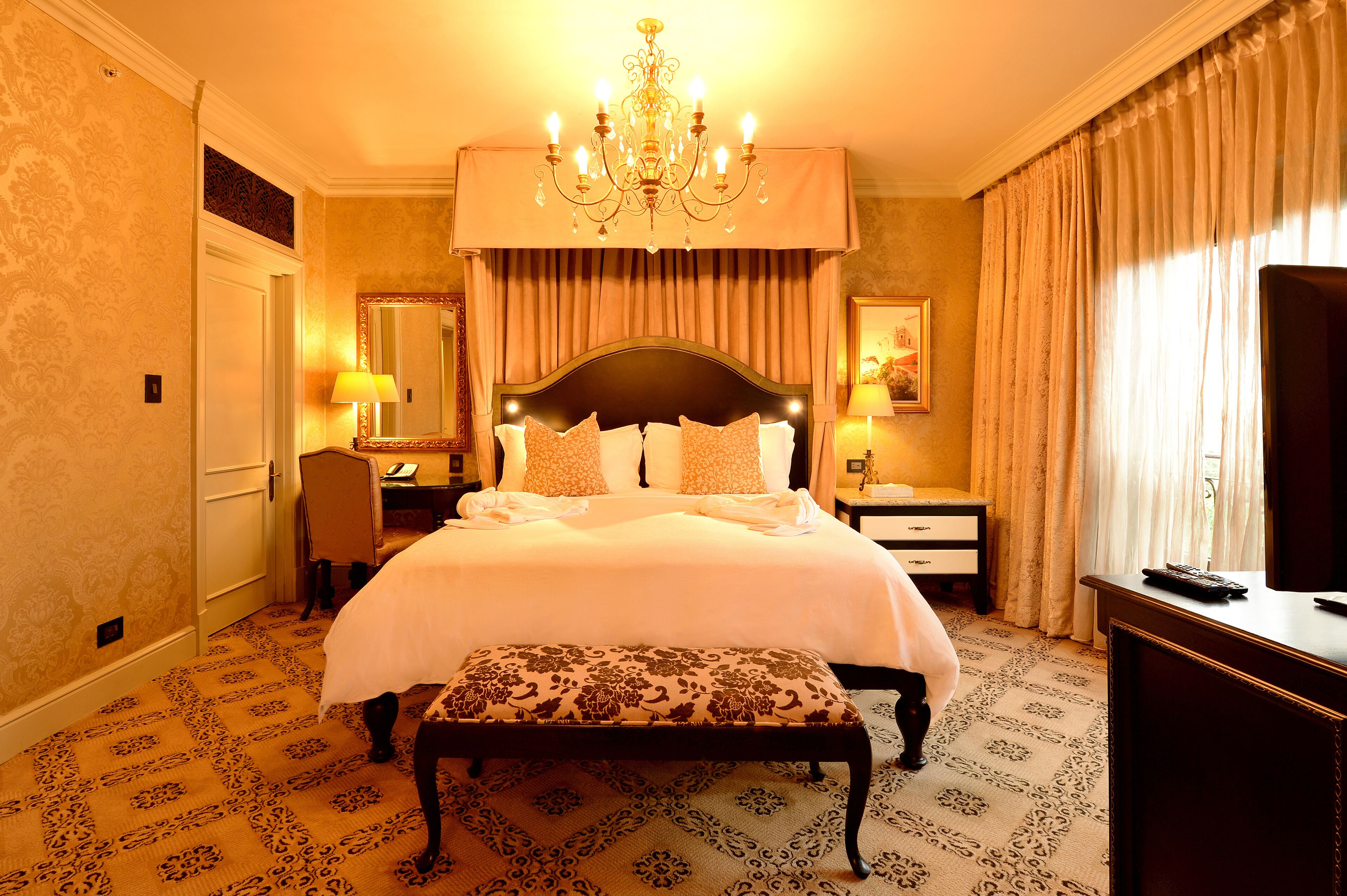 Une chambre d’hôtel avec un grand lit, une banquette, un bureau, une chaise, une télévision et un lustre.
