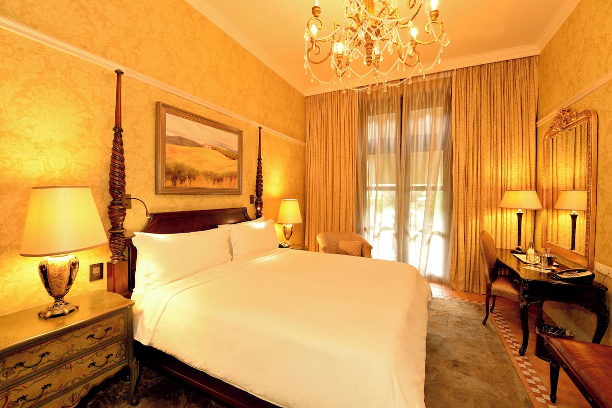 A hotel room with a large bed, two bedside tables with lamps, a desk with a chair, a mirror, and a chandelier.