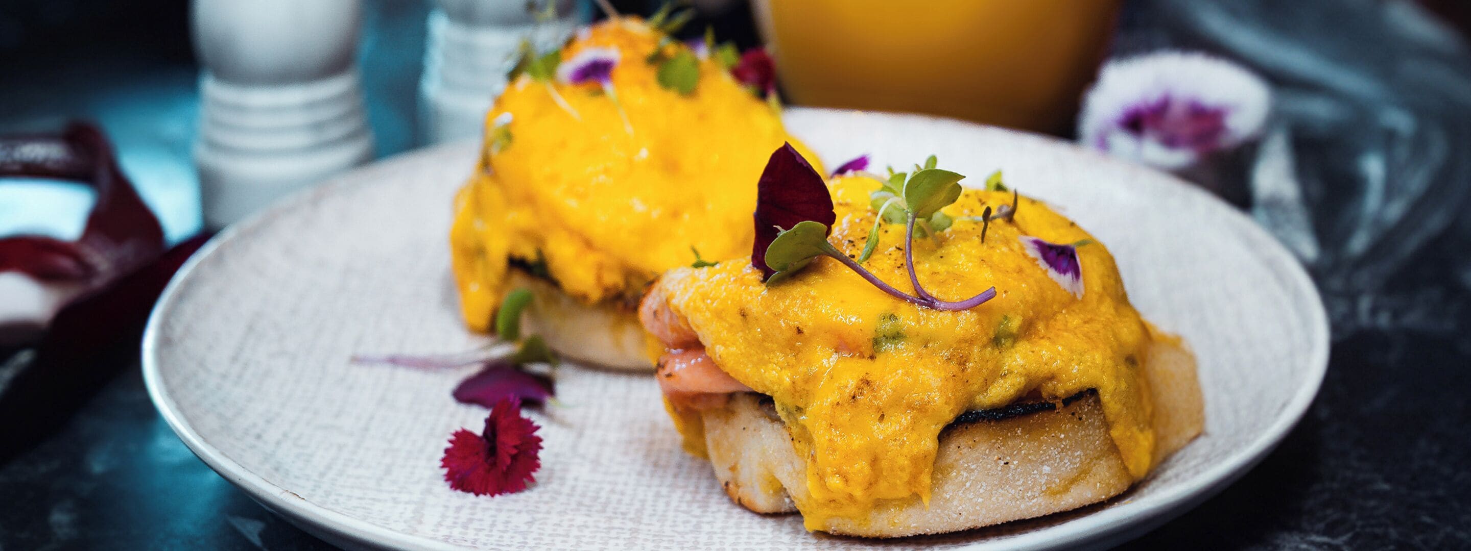 Ein Teller mit einem Frühstücksgericht, möglicherweise Eggs Benedict, garniert mit Kräutern und essbaren Blüten.