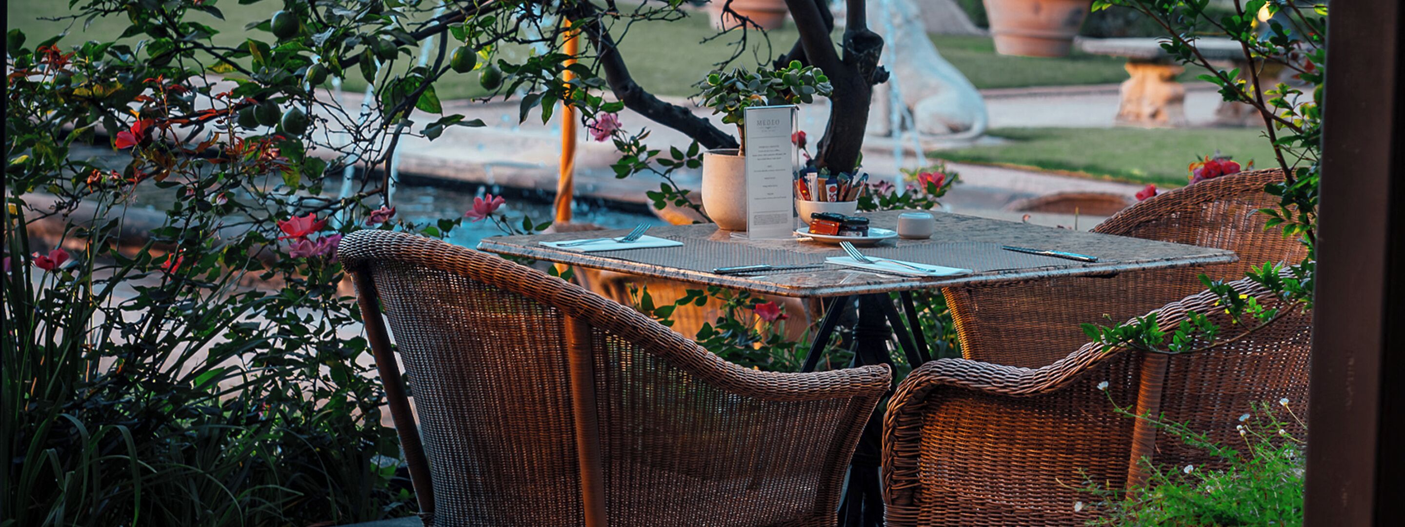 An outdoor dining area with wicker chairs and a table set for two, surrounded by greenery and a small fountain.